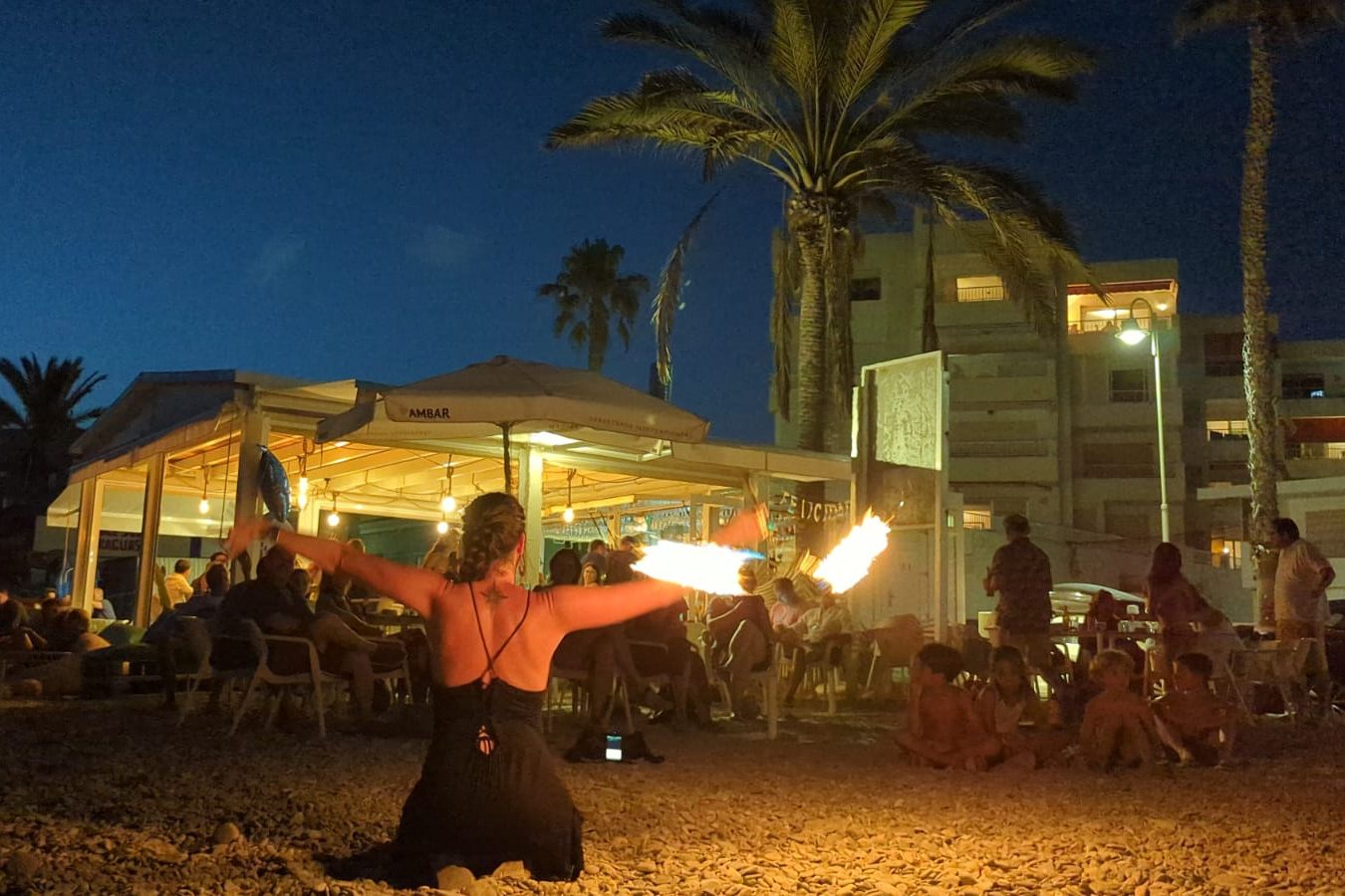 Maka bailando con las dos cariocas de fuego en una mano y de rodillas en el la arena de la playa. La primera línea del público son los niños y niñas invitados al cumpleaños de Olivia